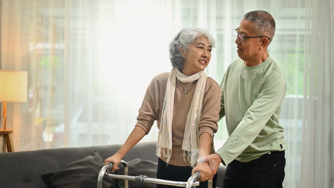 Woman with a walker standing up in a living room with a man helping behind her.