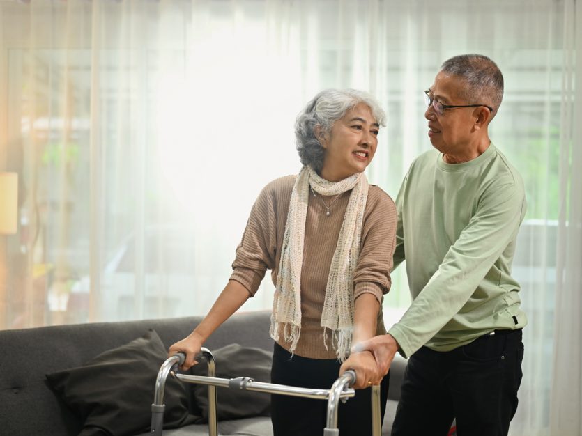Woman with a walker standing up in a living room with a man helping behind her.