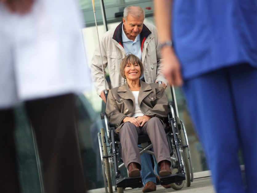 Older man pushing smiling older woman out of the hospital in wheelchair.