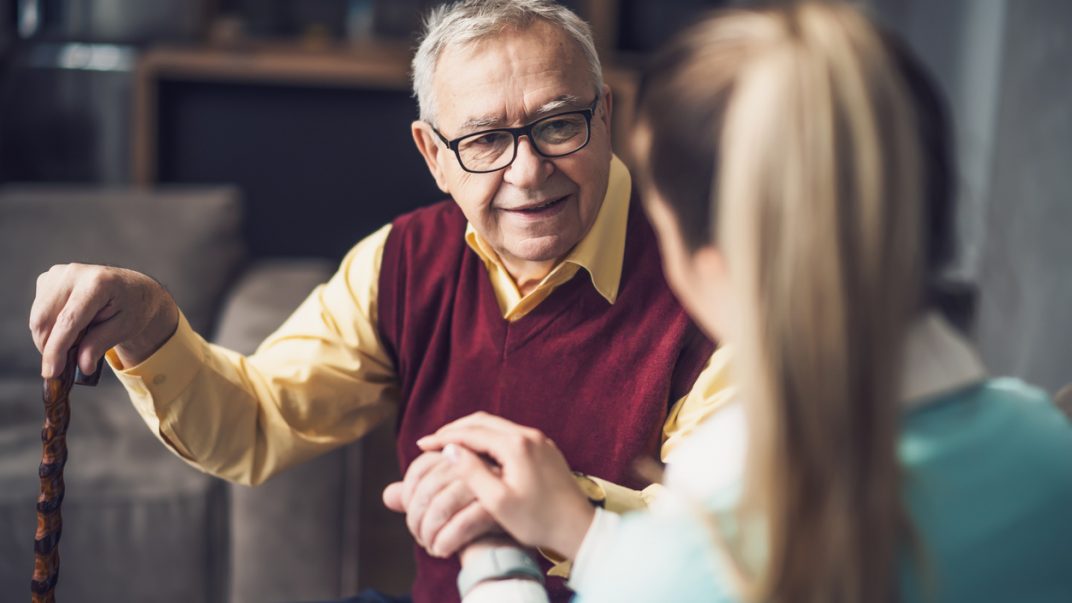 Caregiver holding the hand of an elderly man sitting on a couch with a cane.