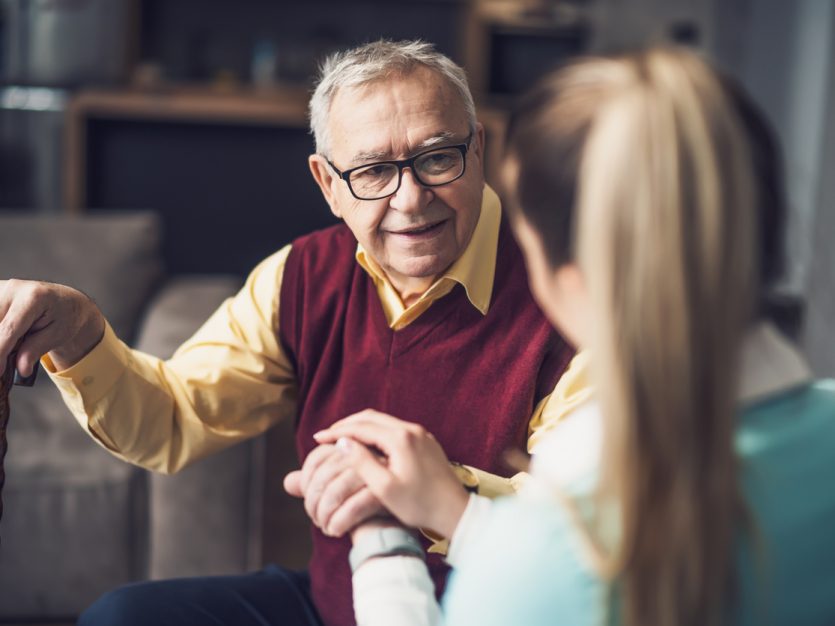 Caregiver holding the hand of an elderly man sitting on a couch with a cane.