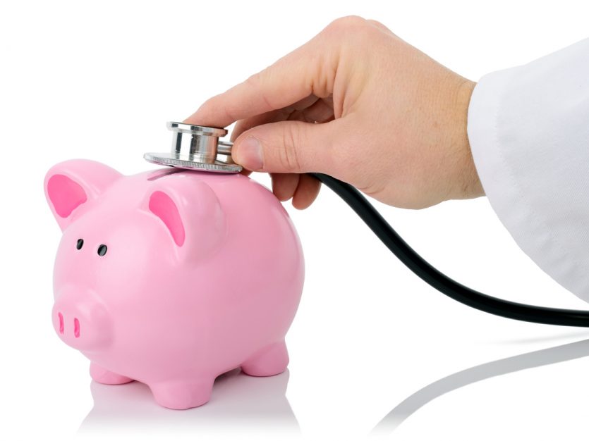 Hand placing a doctors stethoscope on top of a pink piggybank.