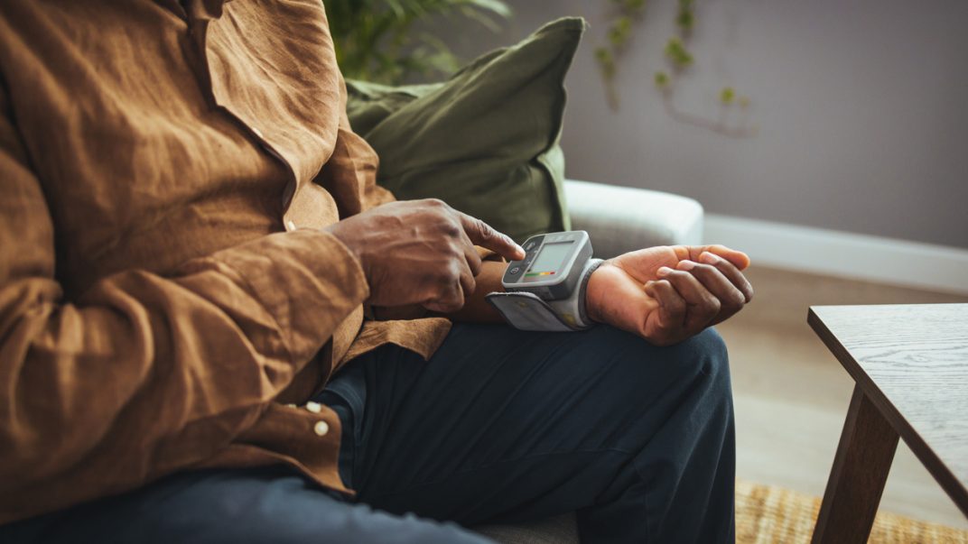 Older adult male sitting at home and using a wrist blood pressure monitor.