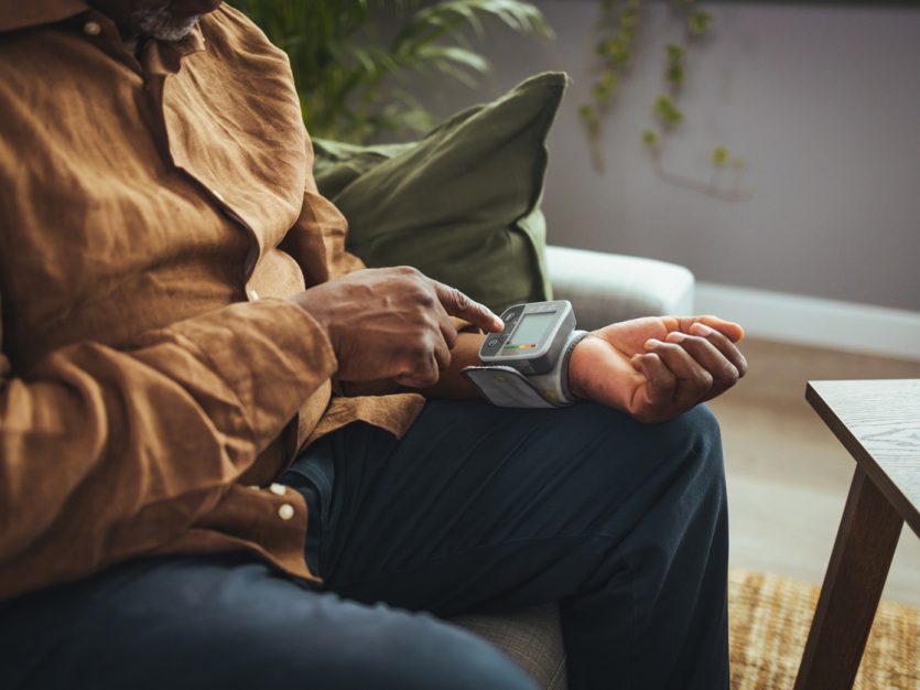 Older adult male sitting at home and using a wrist blood pressure monitor.