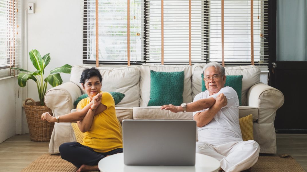 Older adult couple sitting on their living room floor and stretching while look at an open laptop.