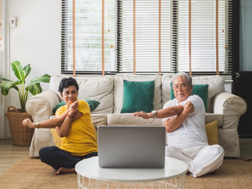 Older adult couple sitting on their living room floor and stretching while look at an open laptop.