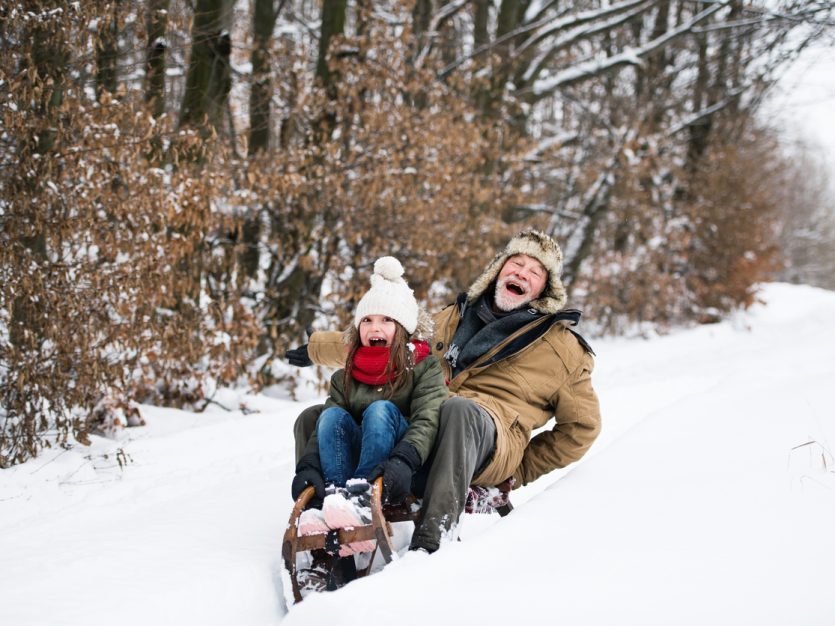 Senior grandfather and young granddaughter sledding and smiling on a winter day.