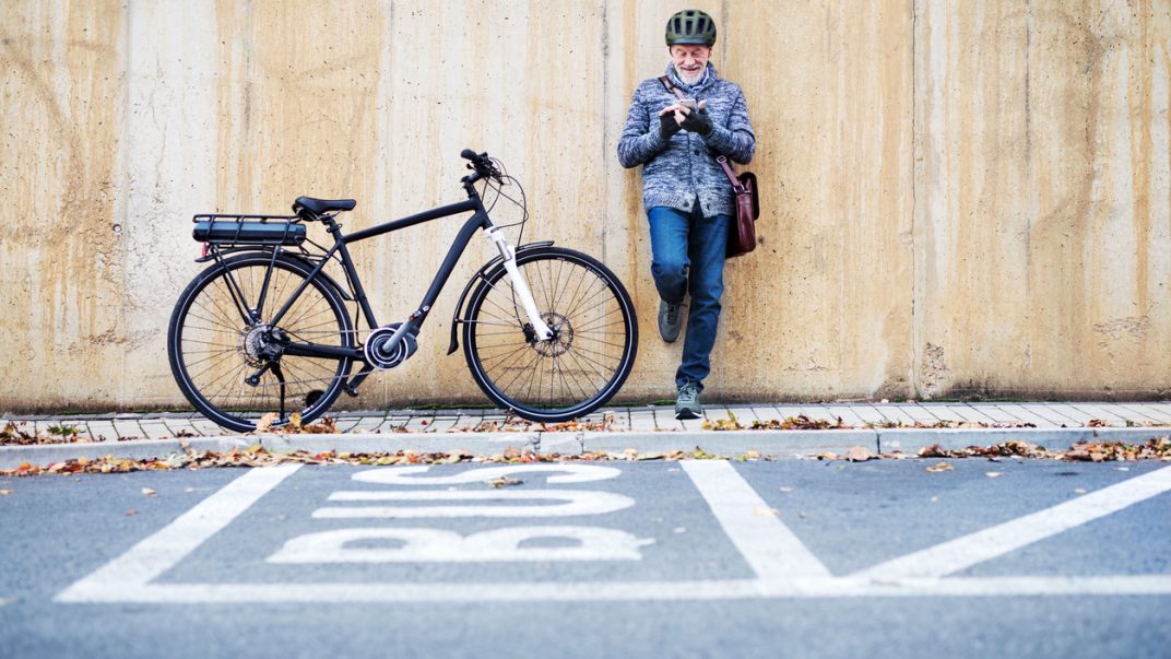 Active senior man leaning against a concrete wall and using a smartphone with his bike next to him.