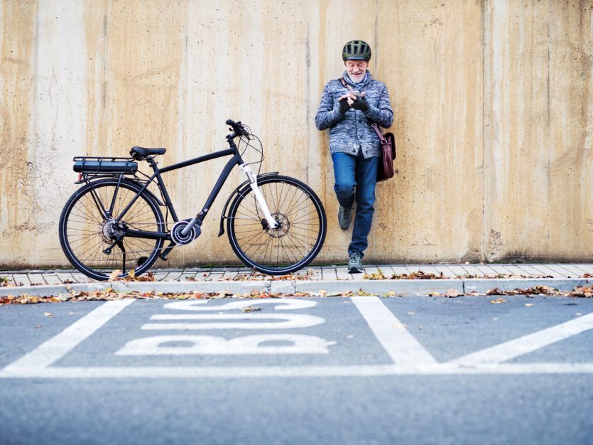 Active senior man leaning against a concrete wall and using a smartphone with his bike next to him.