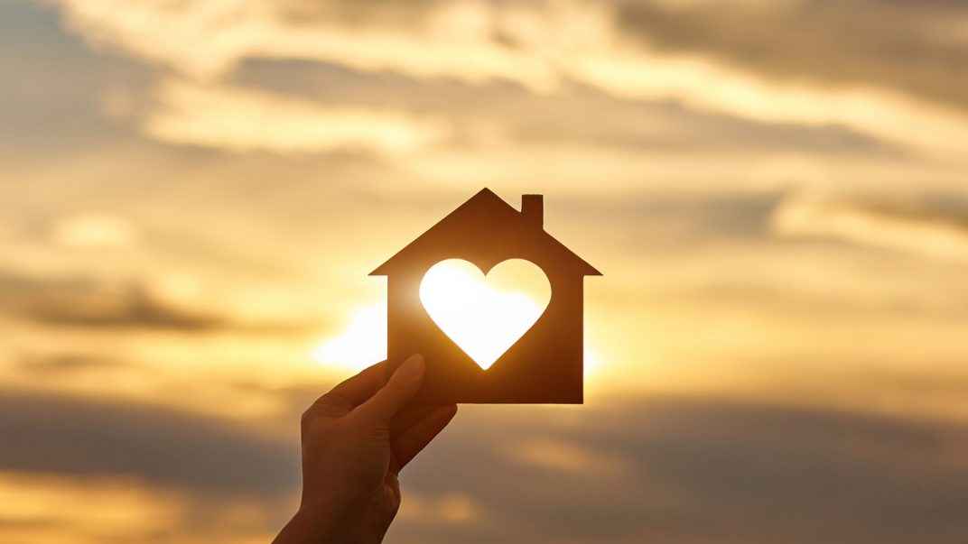 Hand holding a house cut-out with a hole in the form of heart against the sun.