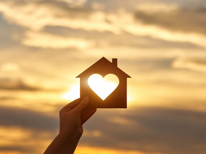 Hand holding a house cut-out with a hole in the form of heart against the sun.