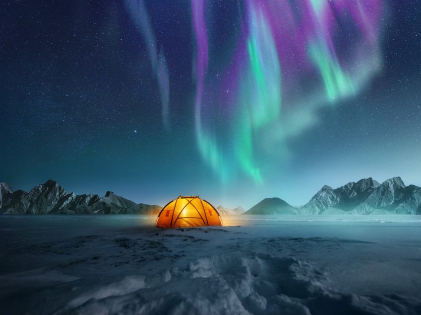 A glowing tent pitched up in the snow at night with the northern lights flickering in the sky above.