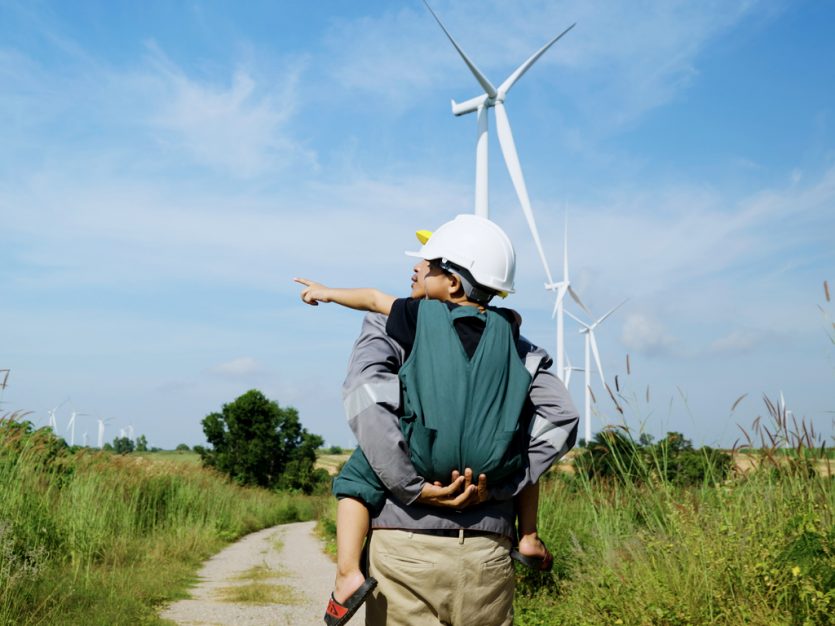Father giving his son a piggyback in a wind turbine field, both wearing hardhats.
