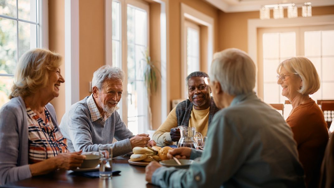 Group of five older adults sitting around a dining room table eating and chatting.