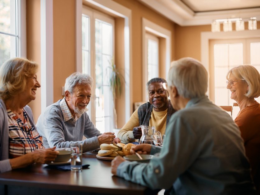 Group of five older adults sitting around a dining room table eating and chatting.