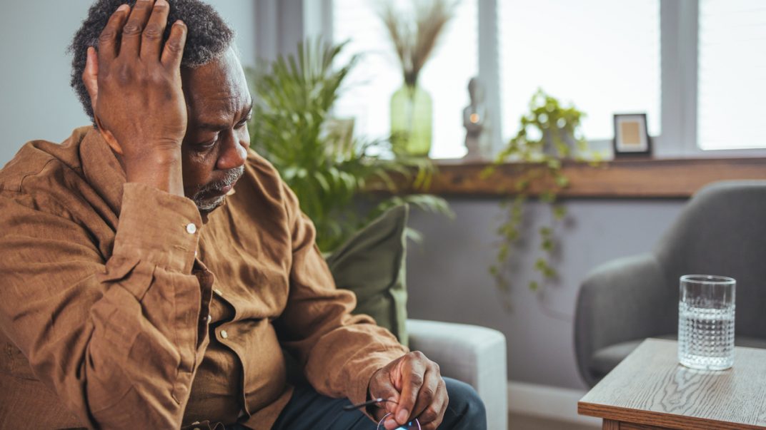 Older adult male sitting on a couch with glasses in one hand and touching his head with the other hand.