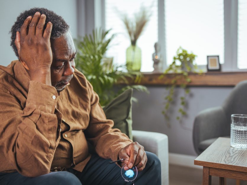 Older adult male sitting on a couch with glasses in one hand and touching his head with the other hand.