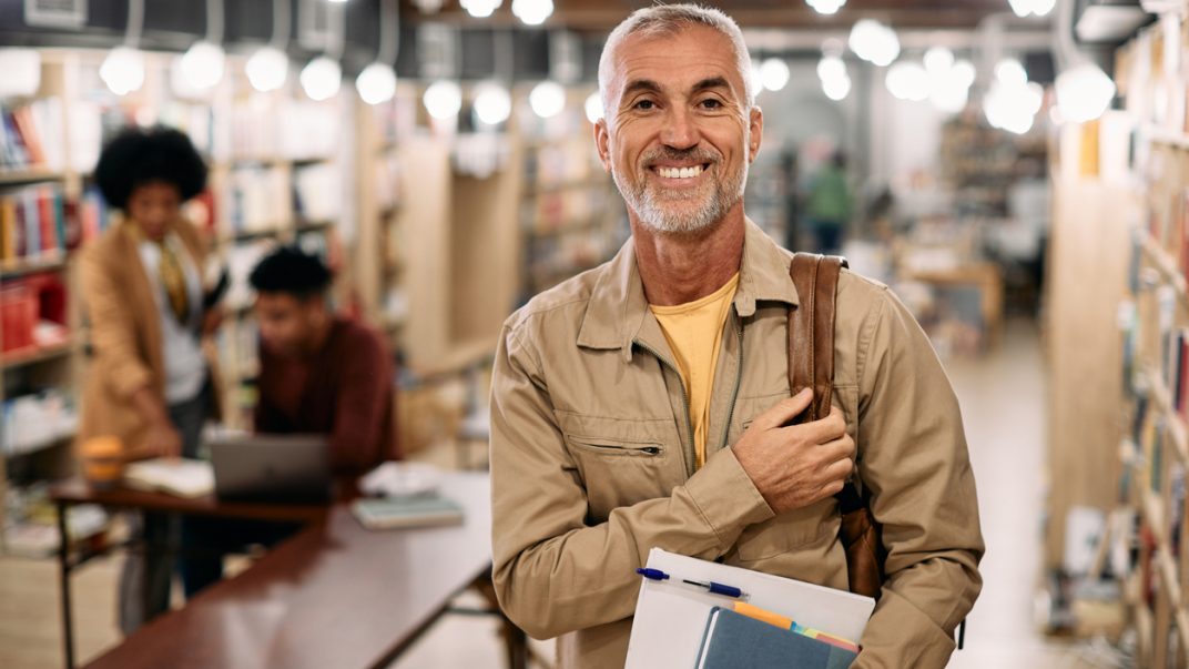 Seniors on the campus 1 Happy mature male student standing in a university library carrying books and looking at camera