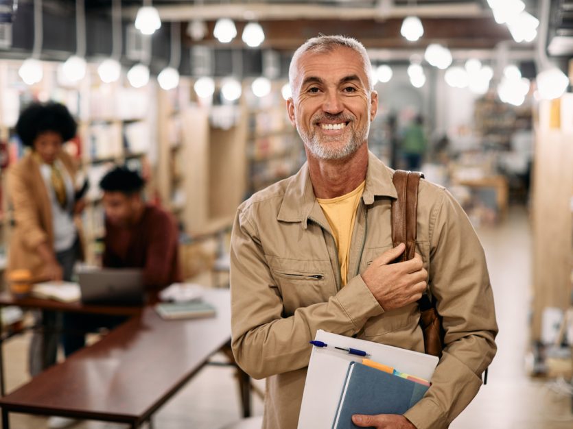 Happy mature male student standing in a university library carrying books and looking at camera