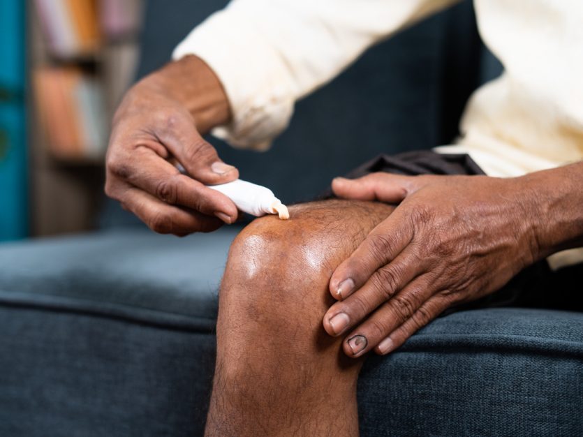 Older adult sitting on a couch and applying ointment to their knee.