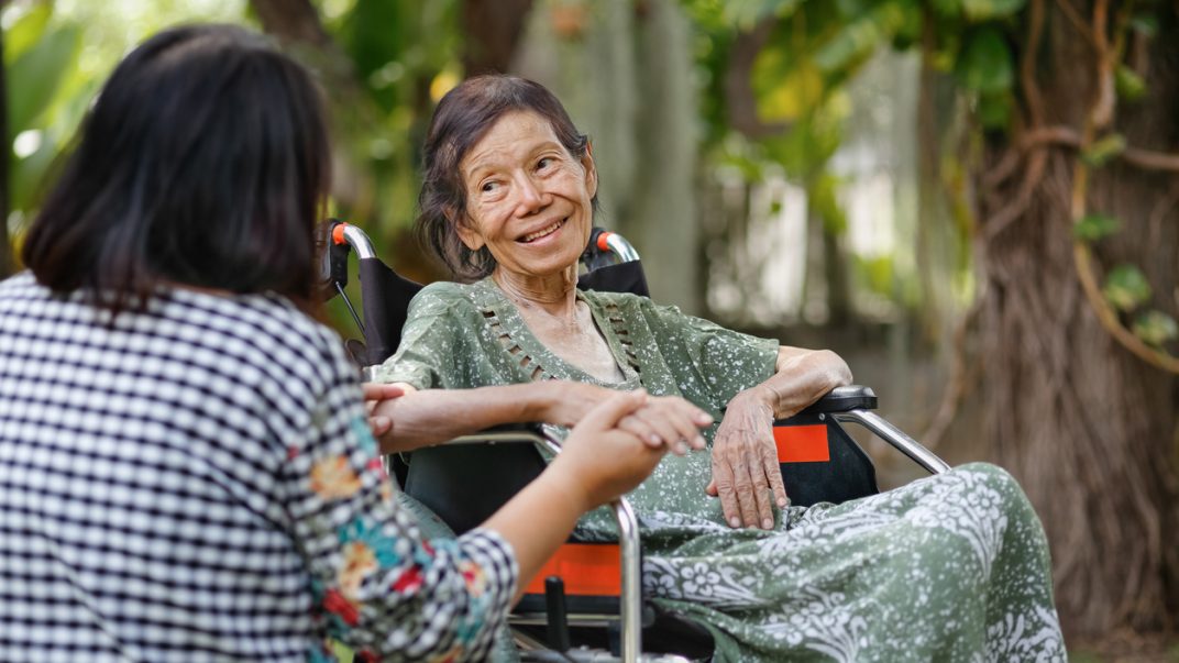 Older adult in a wheelchair sitting outside and talking to a peer who is holding their hand.
