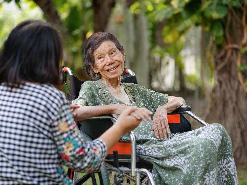 Older adult in a wheelchair sitting outside and talking to a peer who is holding their hand.