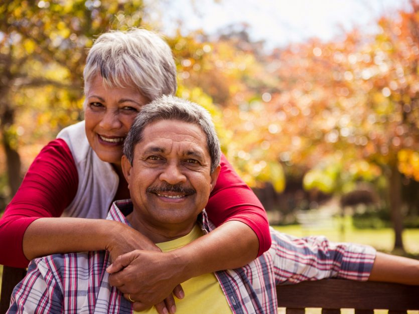 Older adult male sitting on a park bench on a sunny day, while his wife stands behind the bench and hugs him.