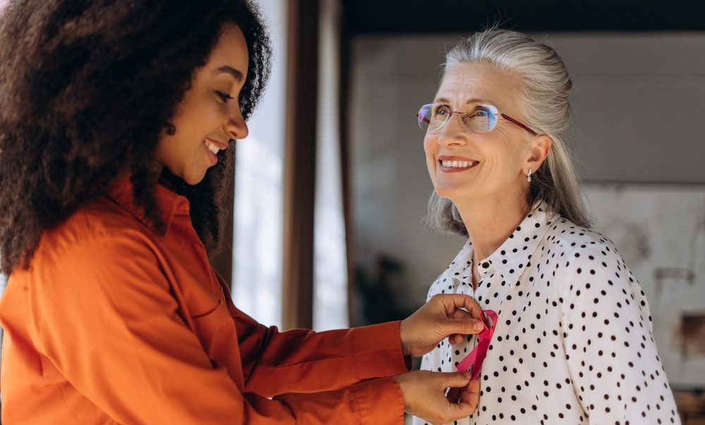 Breast Cancer Awareness Month 2025: Early detection, symptoms, and support 1 A younger woman attaches a pink Breast Cancer Awareness ribbon to an older woman’s top
