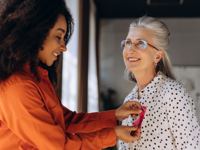 A younger woman attaches a pink Breast Cancer Awareness ribbon to an older woman’s top