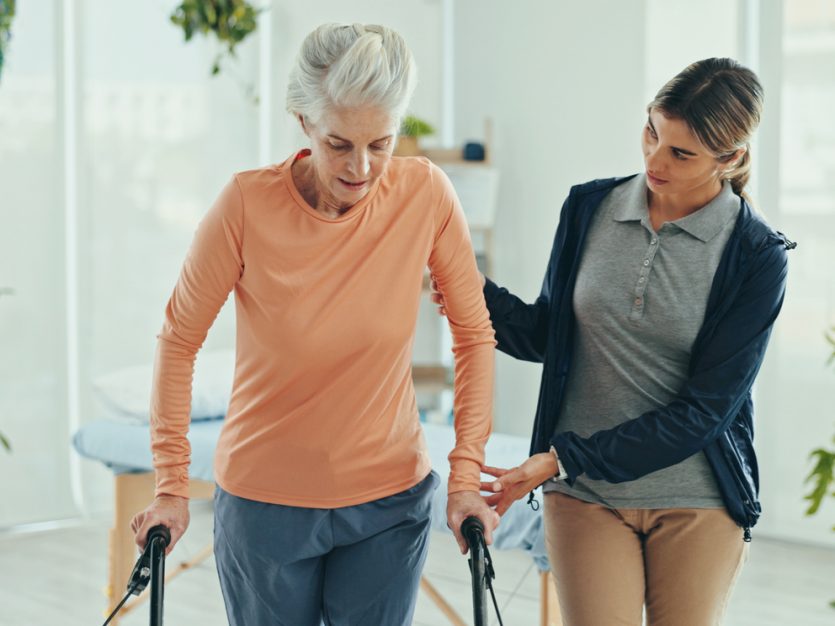 Older adult walking with with the assistance of a physiotherapist and a walking aid.