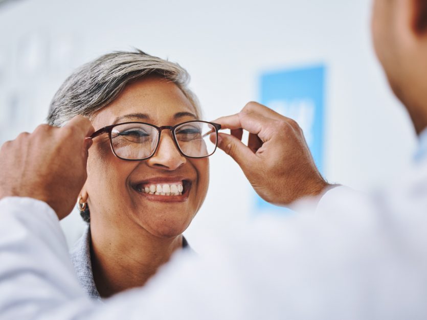 Woman smiling as an eye doctor places eyeglasses on her face.